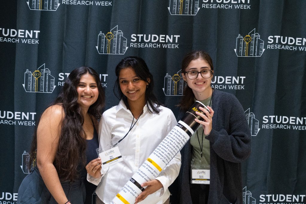 Three friends pose in front of the Student Research Week backdrop