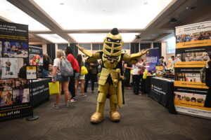 Knightro poses at UCF Grad Fair with his arms outstretched. Around him are tables with advertisements for graduate programs and people talking.