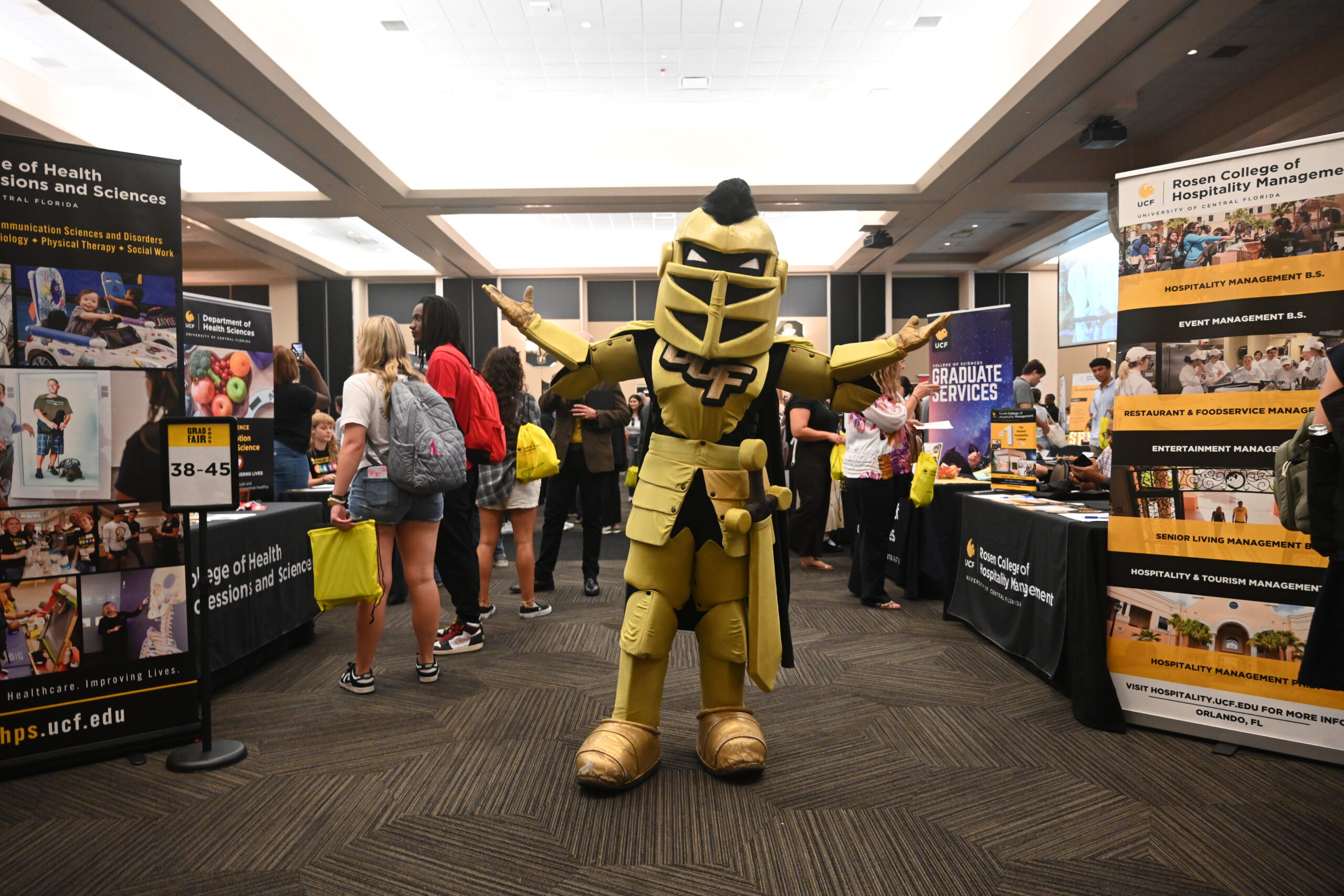 Knightro poses at UCF Grad Fair with his arms outstretched. Around him are tables with advertisements for graduate programs and people talking.